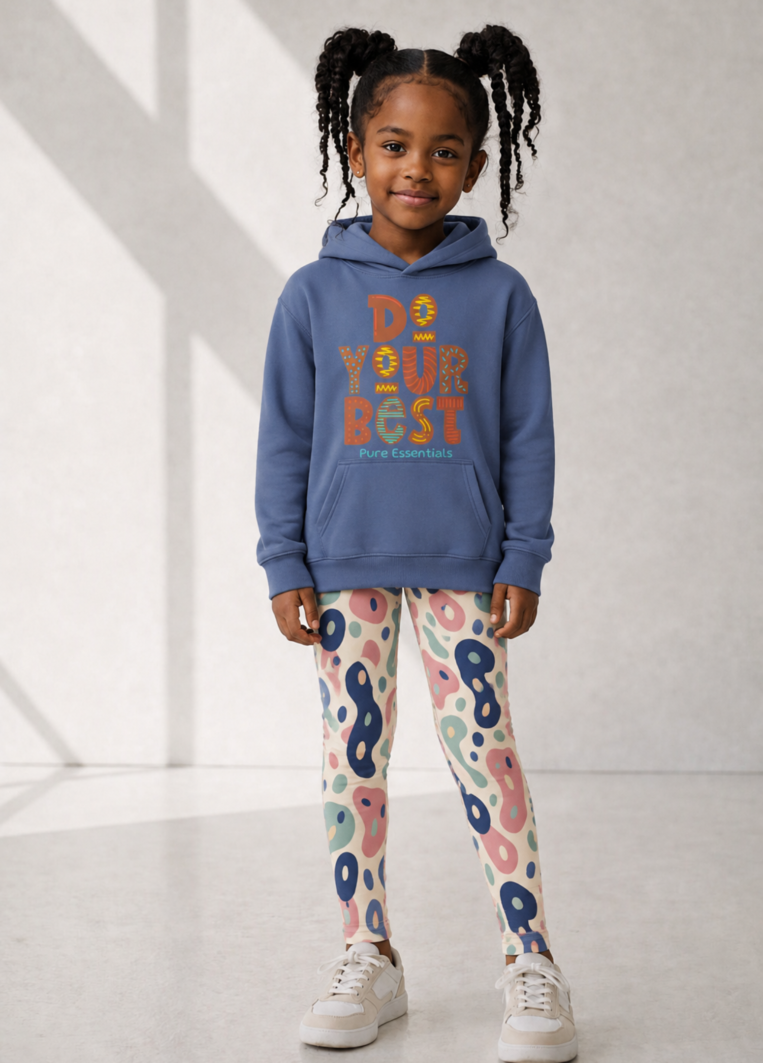 A young girl with braided hair stands in a studio wearing a slate blue hoodie with 'DO YOUR BEST' printed on the front, paired with pink, blue, and white patterned leggings and white sneakers.