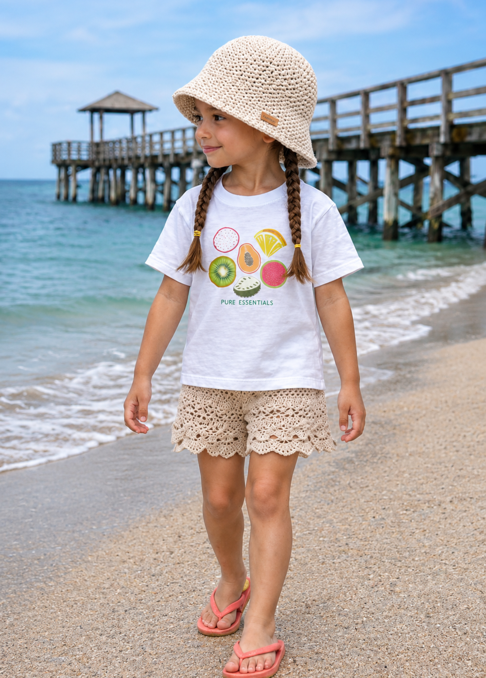 Child on a beach wearing a white t-shirt with colorful design and beige hat.