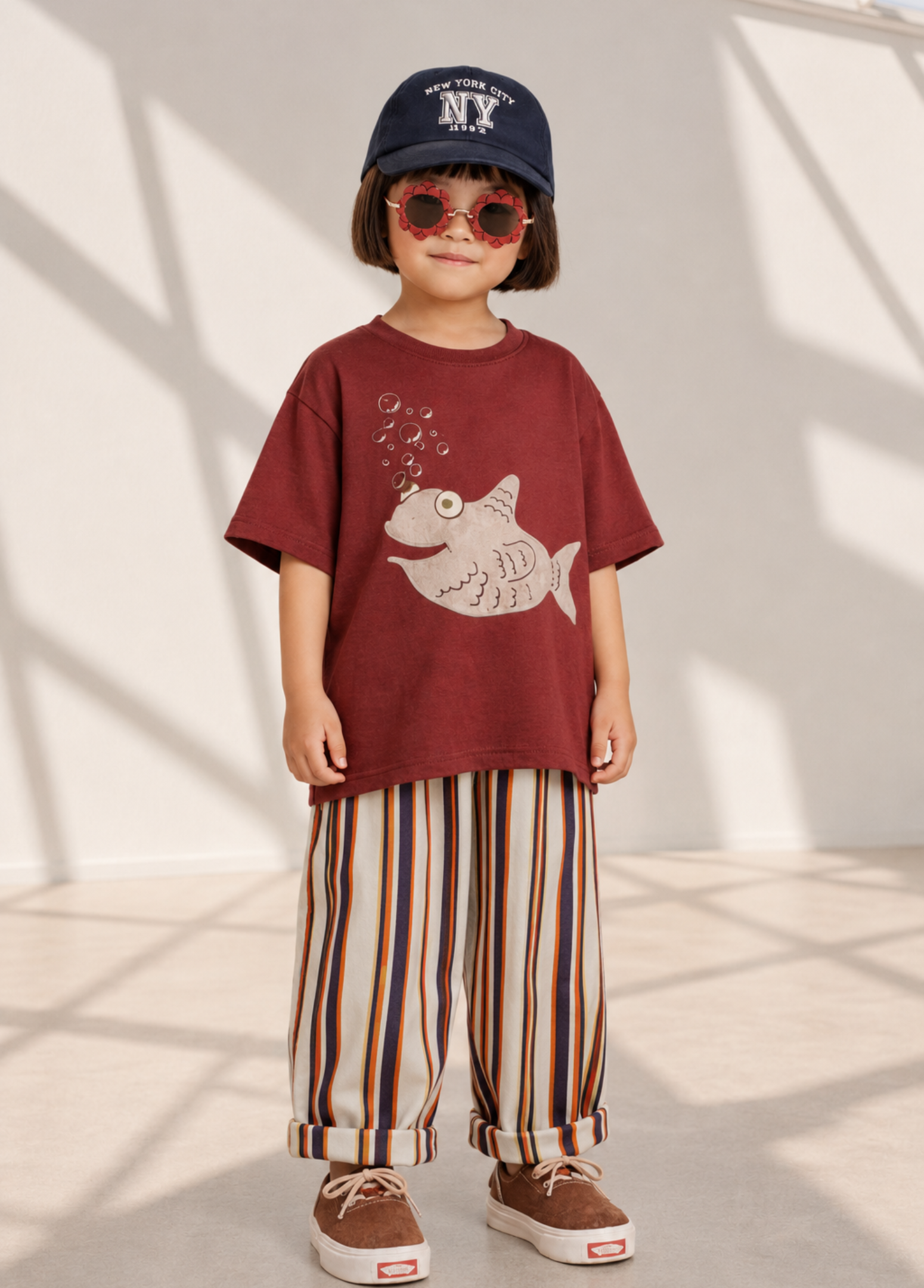 Child wearing a red t-shirt with a fish design, striped pants, and a blue cap indoors.