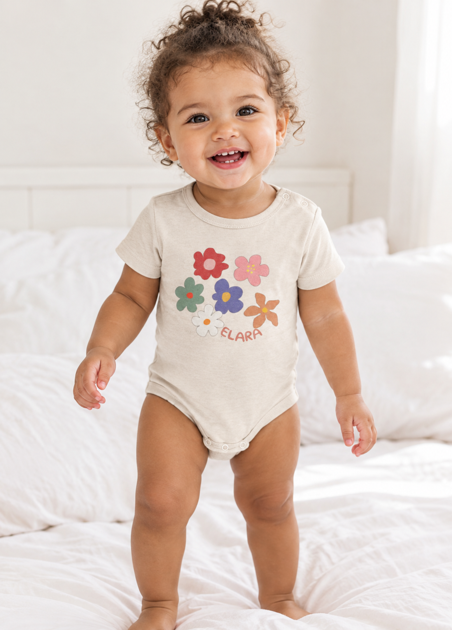 A smiling toddler stands on a white bed wearing a cream short-sleeve bodysuit with colorful flower graphics and the name 'CLAIA' printed on the front.
