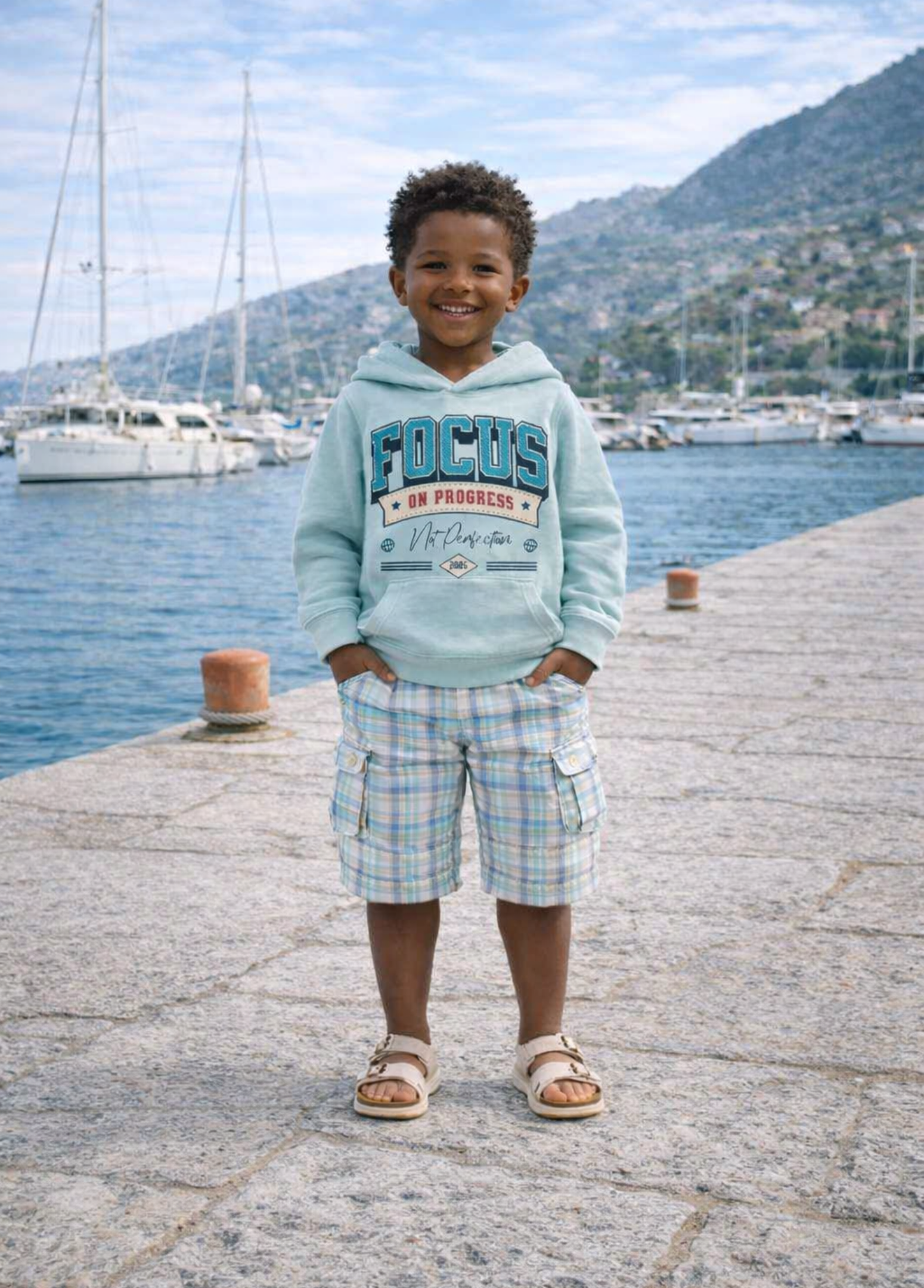 Child wearing a light blue hoodie with text and plaid shorts standing on a dock by a body of water.