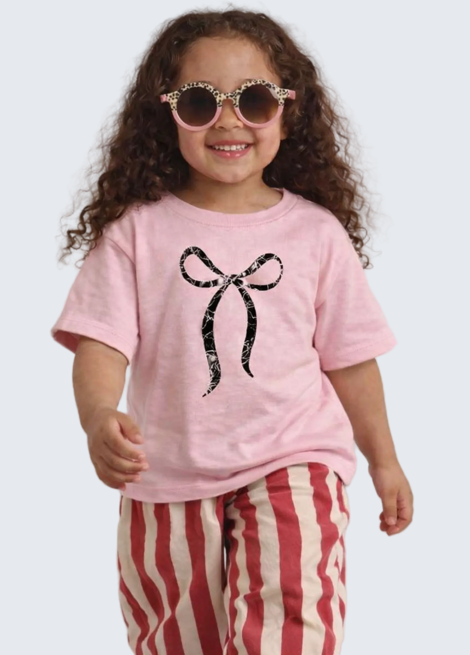 Child wearing a pink t-shirt with a black bow design and red and white striped pants on a light gray background.