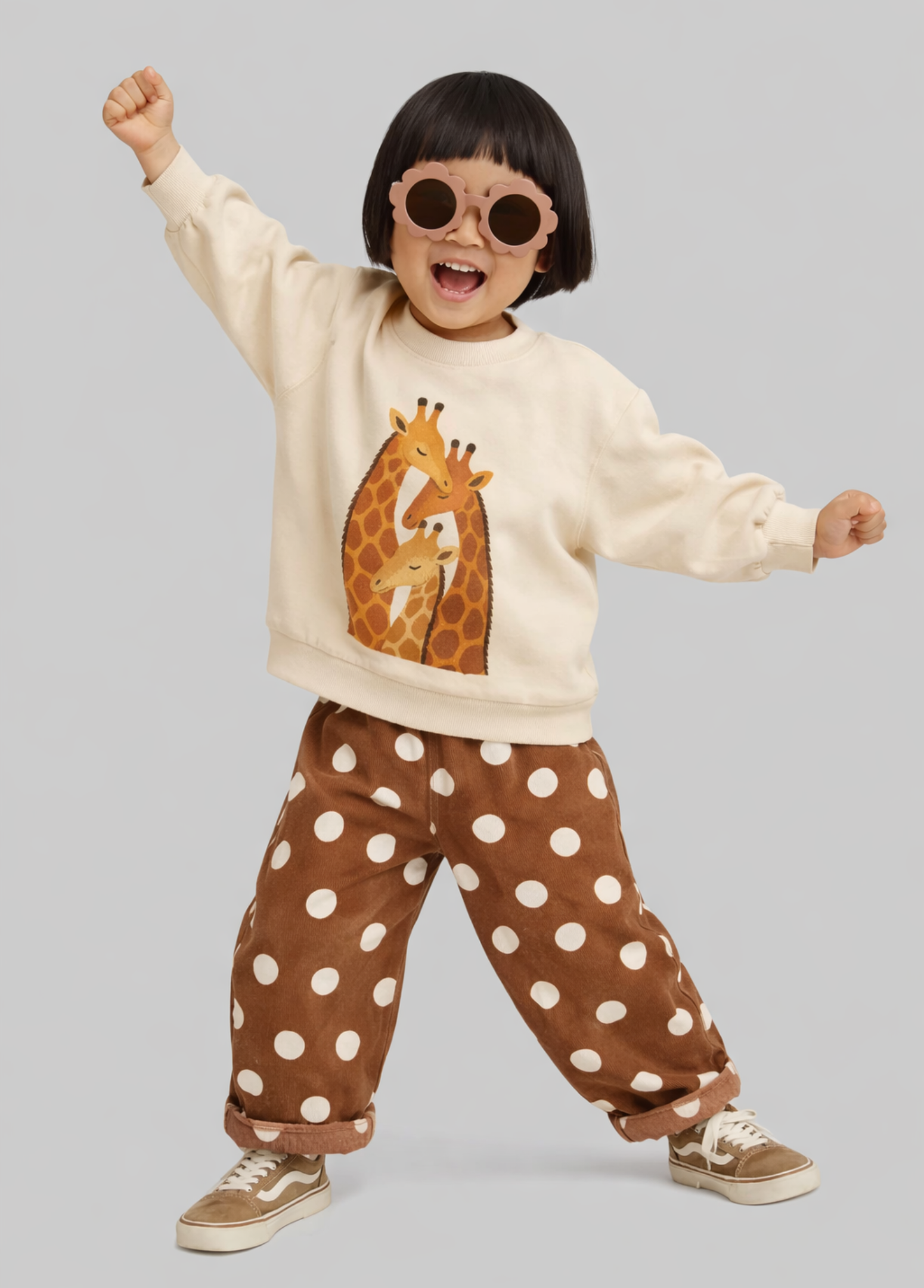 A toddler modeling a cream sweatshirt with a giraffe graphic and brown polka-dot pants, styled with round sunglasses and beige sneakers against a solid gray background.