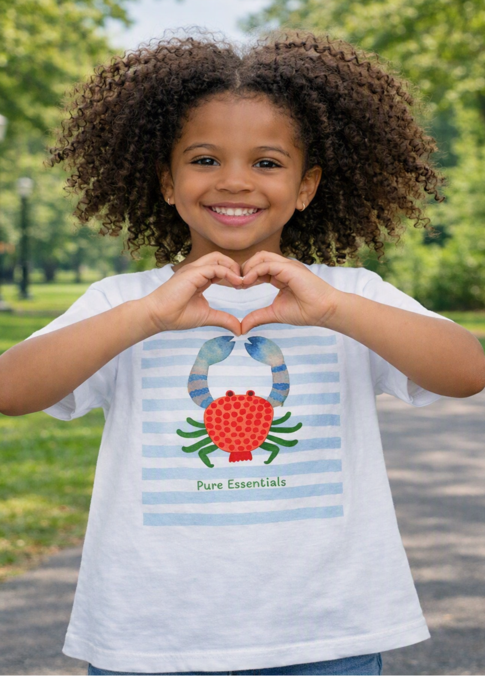 A young girl with curly brown hair stands outdoors in a park, smiling and making a heart shape with her hands. She is wearing a white short-sleeve T-shirt with light blue horizontal stripes and a graphic of a red strawberry and blue birds above the words 'Pure Essentials'.