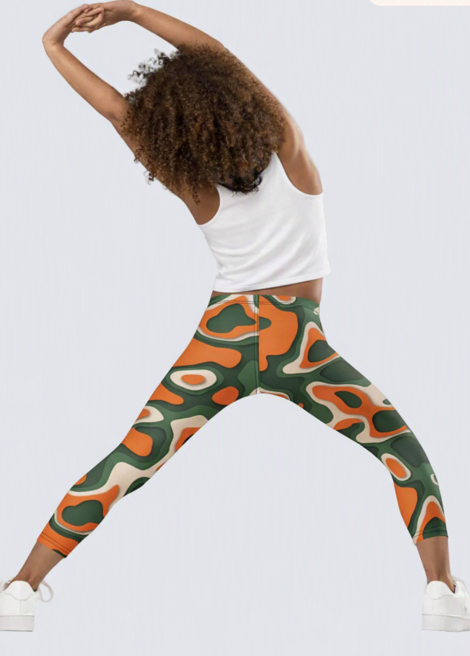 A model wearing vibrant abstract pattern leggings in green, orange, and beige, paired with a white tank top and white sneakers, posing in a side-stretch position against a light gray background.