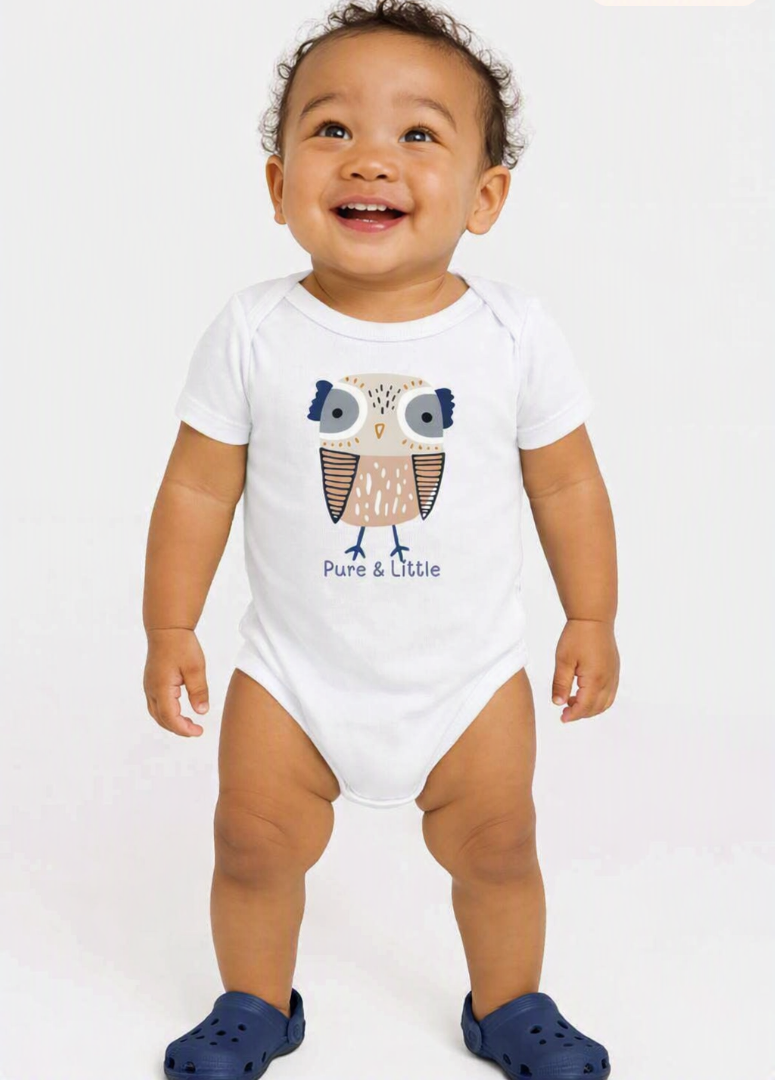 A smiling baby wearing a white short-sleeve bodysuit with a cartoon owl graphic and the text 'Pure & Little' on the front, standing against a light gray background. The baby is also wearing blue clogs, and a 'Made with AI' watermark is visible in the top right corner.