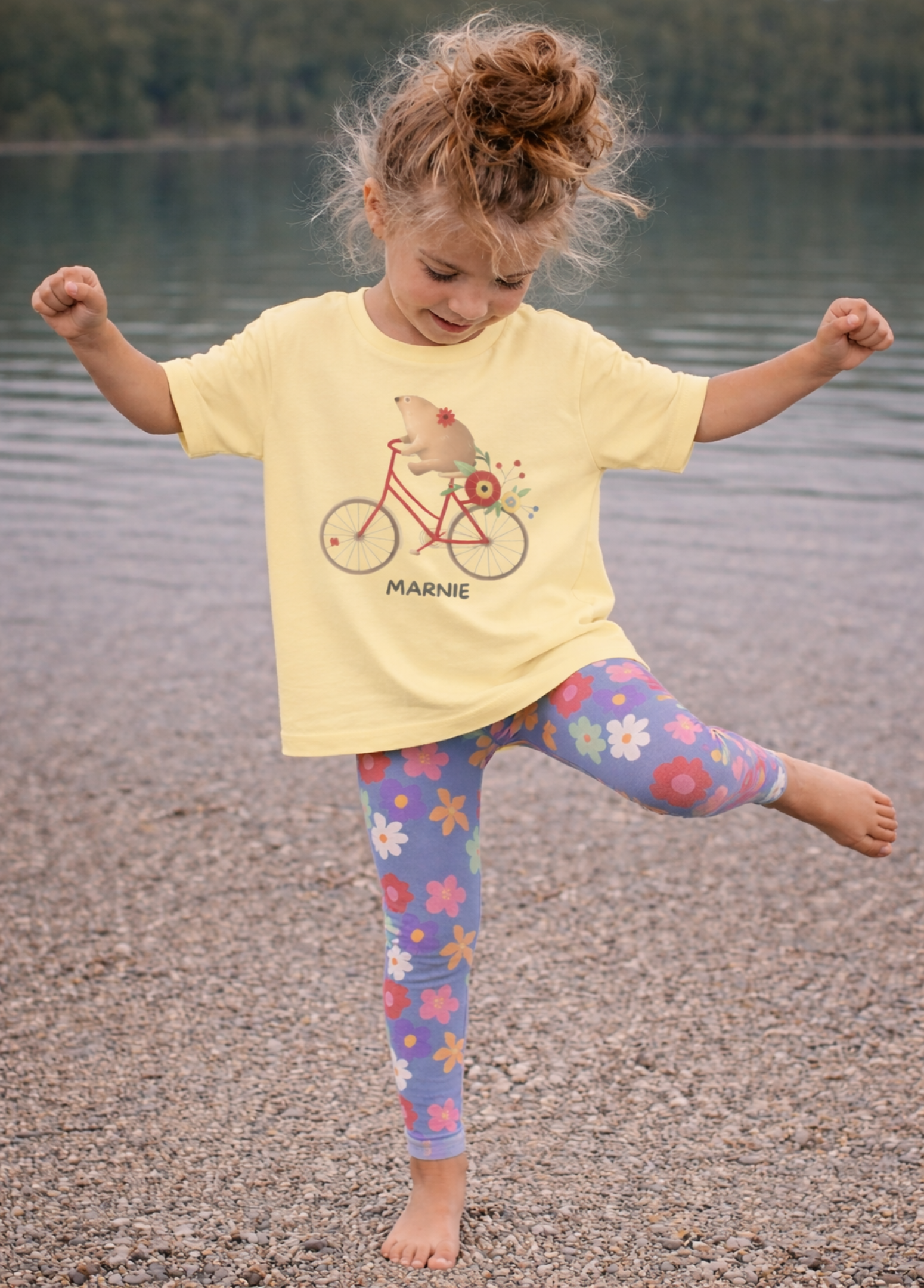 Child wearing a yellow t-shirt with a bicycle graphic and colorful leggings by a body of water.
