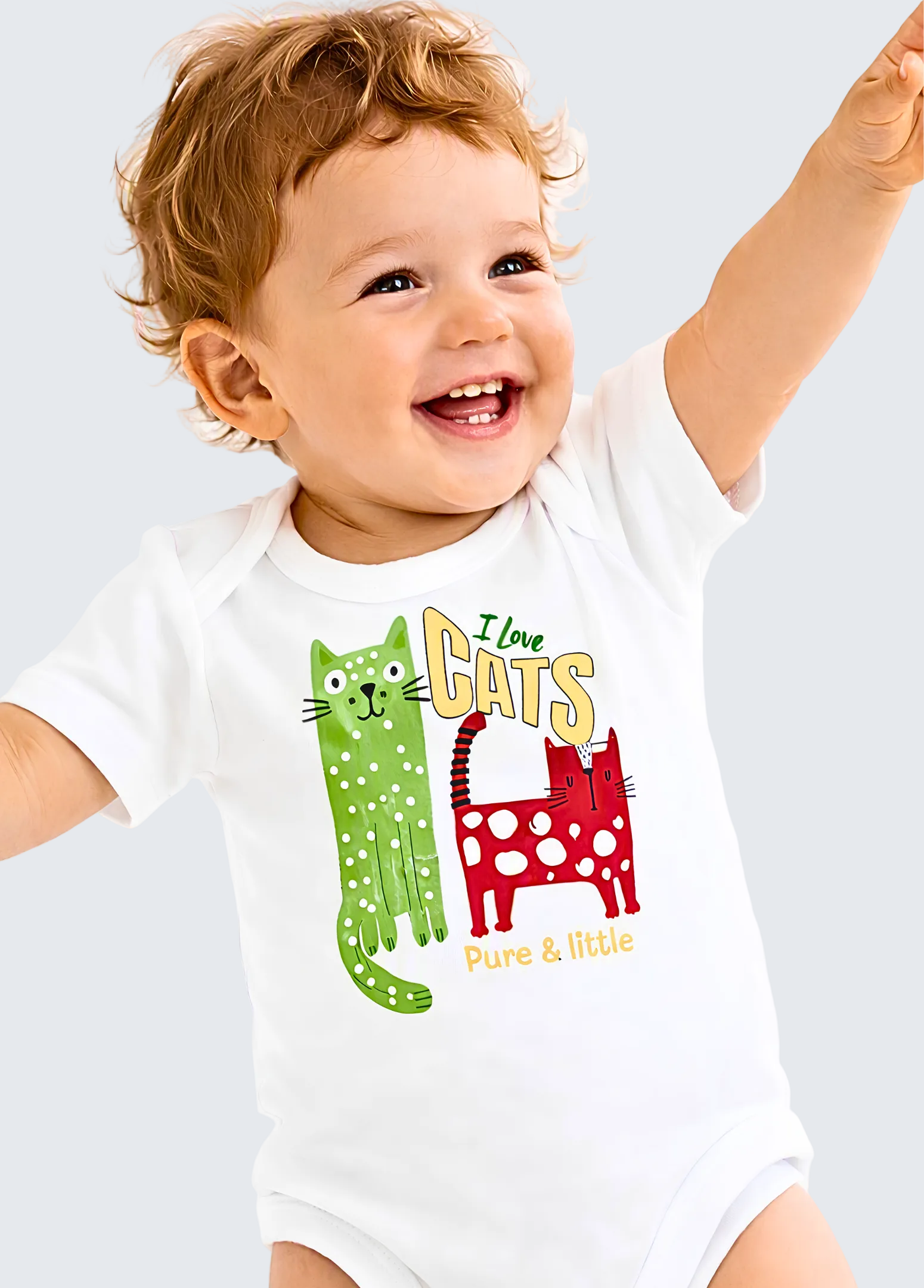 A smiling toddler with curly hair wearing a white short-sleeve bodysuit with a colorful graphic that reads 'I love CATS Pure & little' with two cartoon cats, one green and one red, on a plain white background.