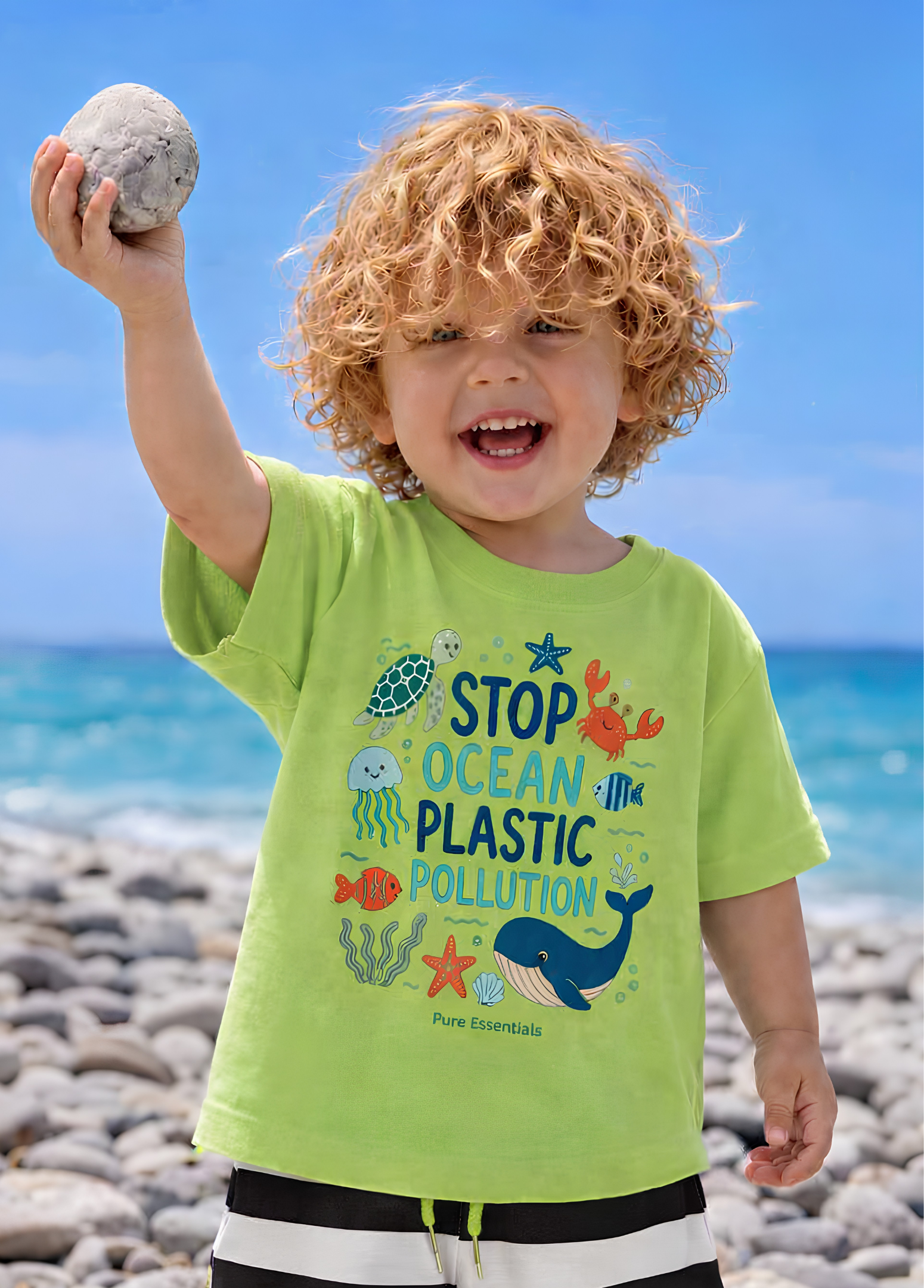 A smiling young child with curly blond hair stands on a rocky beach holding a stone above their head, wearing a bright lime green graphic T-shirt that reads 'STOP OCEAN PLASTIC POLLUTION' with ocean-themed illustrations. The background shows blue sky and ocean water.