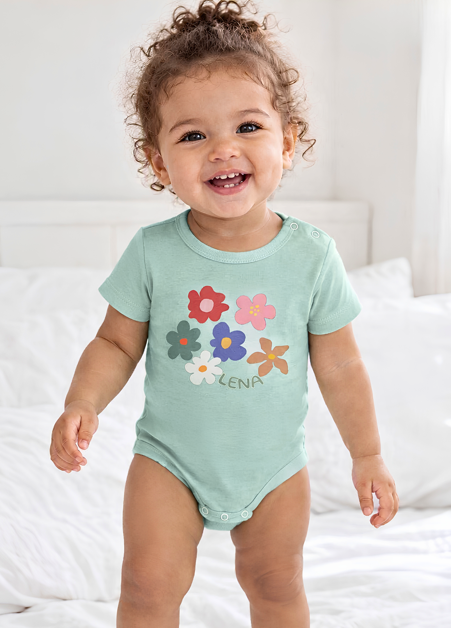 A smiling toddler wearing a mint green short-sleeve bodysuit with colorful flower graphics and the name 'ALENA' printed on the front, standing on a white bed.
